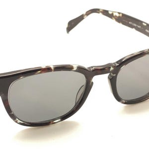 May include: Sunglasses with a dark brown and cream tortoiseshell pattern on the frames and arms. The lenses are a solid gray color. The sunglasses are set against a white background.