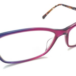 Op de afbeelding: Bril in cat-eye stijl met een gradiënt montuur, van blauw naar magenta. De veren hebben een schildpadpatroon. De bril heeft een zilveren brug en heldere glazen.