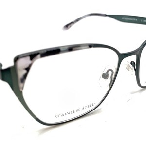 Peut inclure: Lunettes œil-de-chat avec une monture en métal vert foncé et un motif écaille de tortue gris et blanc sur les bords supérieurs. Les verres sont transparents, avec le texte STAINLESS STEEL sur le verre gauche et BCBGMAXAZRIA sur le droit.