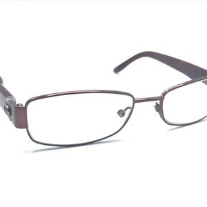 Pode incluir: Um par de óculos de metal cor de vinho com lentes transparentes. Os óculos têm uma forma retangular e uma armação fina de metal.
