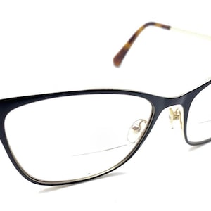 May include: Black cat-eye eyeglasses with gold-tone metal temples and a tortoiseshell pattern on the temple tips. The frame features a small circular gold-tone logo with the letters MK on the outer hinges and clear lenses with a visible bifocal line.