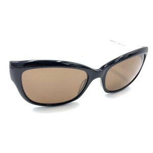 May include: A pair of sunglasses featuring a glossy black plastic frame with a rounded, slightly cat-eye shape. The lenses are a solid, medium-brown tint, providing a clear view through the eyewear. The frame is smooth and uniform in color.