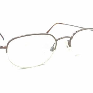Può includere: Un paio di occhiali semi-cerchiati con montatura in metallo color bronzo. Il design presenta aste sottili in metallo con terminali in tartarugato marrone, naselli regolabili e lenti trasparenti fissate da una struttura a filo superiore.