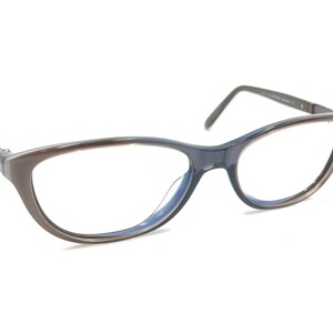 Puede incluir: Gafas de estilo ojo de gato con montura degradada marrón y azul marino. Las gafas tienen lentes transparentes y un diseño elegante y moderno. Las patillas son finas y oscuras, y la estética general es sofisticada.