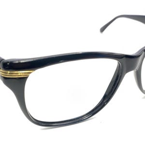 May include: Black cat-eye eyeglasses with gold-colored accents on the temples. The frames have a glossy finish and a classic, elegant design. The lenses are clear, and the arms curve slightly for a comfortable fit.