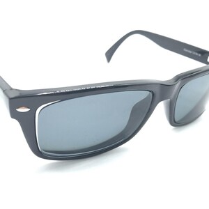 May include: Black sunglasses with a rectangular frame and dark grey lenses. The frame features a silver-toned accent on the outer corners near the hinges. The temples are black and connect to the frame with a visible hinge mechanism.