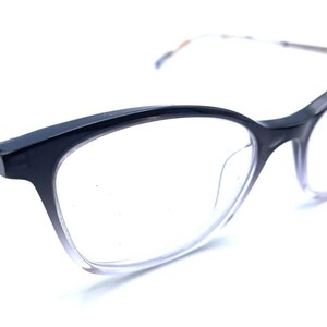 May include: Eyeglasses featuring a dark navy blue frame that transitions into a clear, transparent lower rim. The temples are thin and metallic, showcasing a light gold or bronze tone, providing a sleek contrast to the bold upper frame.