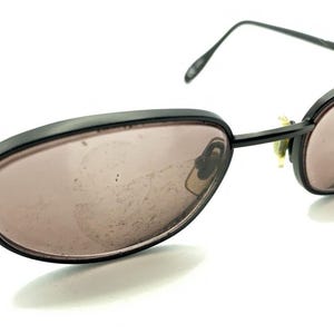 Può includere: Occhiali da sole con montatura in metallo nero e lenti ovali. Le lenti sono colorate di un marrone chiaro.