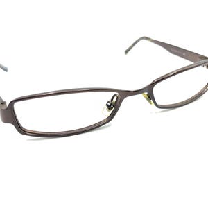 May include: Brown rectangular eyeglasses with a thin metal frame. The glasses have a classic design with a subtle curve at the top of the lenses. The temples are thin and extend to the ear pieces.