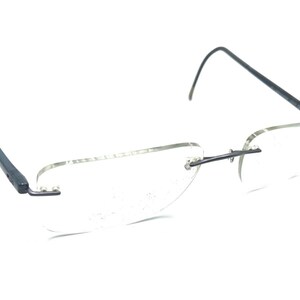 May include: Rimless eyeglasses featuring clear lenses and thin, dark charcoal metal temples. The frame design includes a bridge and hinges that connect directly to the lenses, showcasing a sleek, lightweight construction with curved temple tips.