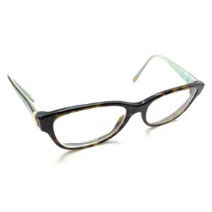 May include: Eyeglasses featuring a dark brown tortoiseshell pattern on the front frames. The temples are a light mint green color, connected to the front by a small gold-toned metal accent. The lenses are clear and the frame shape is slightly cat-eye.