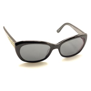 May include: Black sunglasses with a cat-eye frame shape and dark gray lenses. The temples feature a series of small, metallic gold-toned studs embedded along the side, providing a decorative accent to the otherwise solid black plastic frame design.
