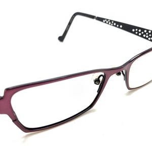May include: Eyeglasses with a deep purple rectangular frame and black temples. The temples feature a decorative pattern of small, circular cutouts along the sides. The nose pads are clear, and the lenses are transparent and clear.