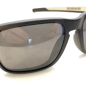 Puede incluir: Gafas de sol gris oscuro mate con lentes oscuras reflectantes y dos pequeños detalles circulares de color verde lima en las esquinas exteriores de la montura. Las patillas presentan un interior beige claro con el texto HOLBROOK MIX impreso en negro.