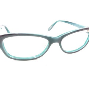 May include: A pair of teal-colored eyeglasses with a cat-eye frame. The glasses have a gradient effect, transitioning from a darker shade at the top to a lighter teal towards the bottom. The lenses are clear, and the arms are thin and teal.
