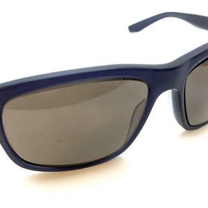 May include: Navy blue sunglasses with brown tinted lenses. The sunglasses have a rectangular frame and a sleek, modern design. The arms of the sunglasses are thin and curve slightly. The lenses are a dark brown color, providing protection from the sun.