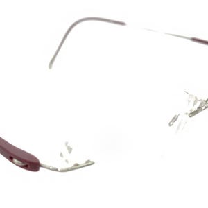 Pode incluir: Óculos sem aro com armação prateada e hastes bordô. Os óculos têm um design minimalista, com foco nas lentes transparentes. Os detalhes bordô adicionam um toque de cor ao design neutro. Os óculos são leves e modernos.