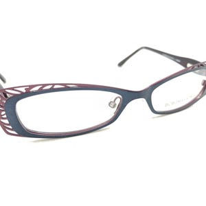 May include: A pair of eyeglasses with a dark blue frame and burgundy accents. The frames have a decorative, openwork design near the temples. The lenses are clear, and the arms are a dark gray color.