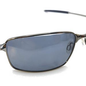 Puede incluir: Gafas de sol rectangulares con montura de metal de cañón de fusil y lentes grises. Las patillas son finas y ligeramente curvadas. Sobre un fondo blanco.