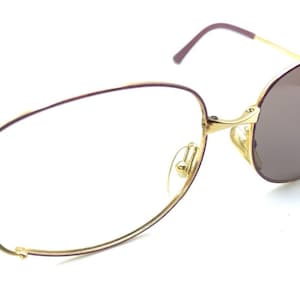 May include: Gold-rimmed spectacles with a distinctive design. The frames have a gold finish with burgundy-coloured temple tips. One lens is a brown tint, while the other is clear. The spectacles are set against a white background.