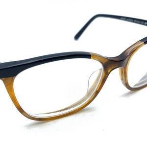 May include: Eyeglasses featuring a two-tone frame with a glossy black upper rim and a translucent amber brown lower rim. The temples are black with a small gold-colored accent near the hinge. The clear lenses are set within a cat-eye shape frame.