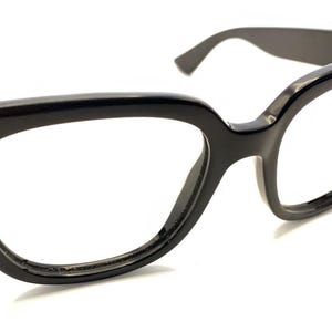 May include: Glossy black cat-eye glasses featuring a thick, bold frame design with wide rims and tapered temples. The eyewear has a distinct angular shape at the outer corners, providing a sharp silhouette. The lenses are clear and transparent.