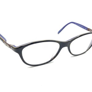 Puede incluir: Gafas negras con forma de ojo de gato. Las monturas tienen un fino borde plateado y detalles azules en las patillas. Las lentes son transparentes y las varillas son de color marrón oscuro.