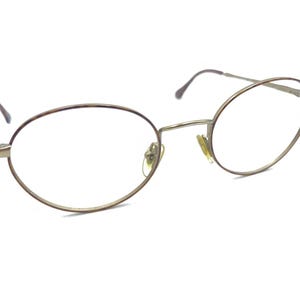 May include: Oval spectacles with a thin, gold-coloured metal frame. The lenses are clear, and the temples are a darker brown. The glasses are set against a white background, highlighting their design and structure.