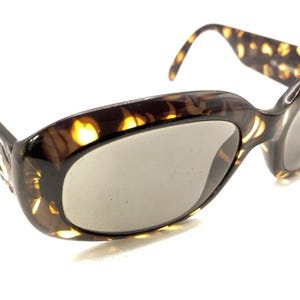 Op de afbeelding: Vintage cat-eye zonnebril met een schildpadpatroon. De monturen zijn bruin en amberkleurig, met grijze glazen. De pootjes hebben een decoratief ontwerp. Een klassiek brillenaccessoire.