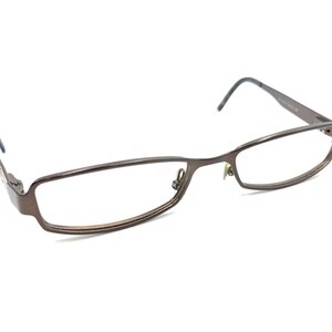 May include: Brown rectangular eyeglasses with a thin metal frame. The glasses have a classic design with a bridge and temples. The lenses are clear, and the overall style is modern and understated.