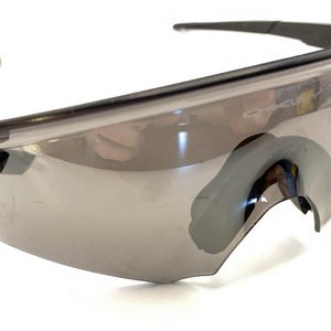 Pode incluir: Óculos de sol esportivos pretos e cinzentos com design envolvente. As lentes tingidas estão em uma armação elegante e moderna. O logotipo da Oakley é visível na haste.