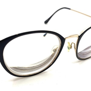 May include: Black and gold-tone spectacles with an oval frame. The frames are black with gold-tone detailing on the temples and bridge. The lenses are clear, and the spectacles are set against a white background.