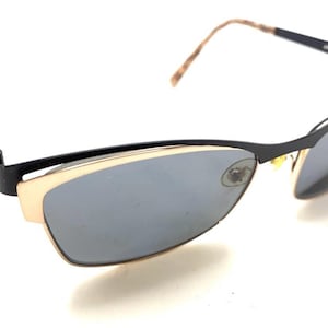 Op de afbeelding: Zwart en goudkleurige zonnebril met grijze glazen. De zonnebril heeft een strak, modern design met een dun zwart montuur en gouden accenten. De veren zijn lichtbruin van kleur. De glazen zijn grijs.