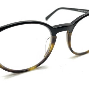 May include: Eyeglasses featuring a two-tone frame design with a solid black upper rim and a brown tortoiseshell pattern on the lower rim. The glasses have thin black temples and clear lenses, showcasing a classic rounded shape with a bridge.