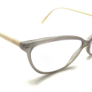 May include: Cat-eye glasses featuring a translucent gray frame with a subtle glitter finish. The temples are a contrasting cream color, and the frame is secured with small silver-toned metal rivets at the outer corners of the front rims.