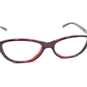 Puede incluir: Gafas con montura burdeos y lentes transparentes. Las monturas de ojo de gato tienen un acento rojo en los bordes interiores. Las patillas son de color gris oscuro. Adecuadas para la corrección de la visión.