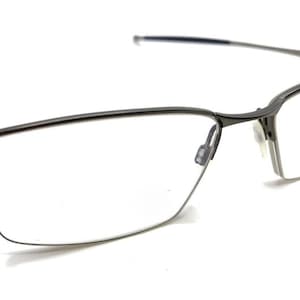 Könnte beinhalten: Eine Brille in Gunmetal-Grau mit einem halbrandlosen, rechteckigen Rahmendesign. Die Brille verfügt über dünne, leichte Metallbügel und verstellbare Nasenpads mit einem glatten, polierten Finish an der Brücke und den oberen Rändern.