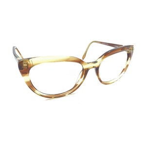 Puede incluir: Gafas de estilo ojo de gato con una montura translúcida de color marrón miel jaspeado y acabado pulido. Las gafas tienen lentes transparentes y varillas finas a juego que se extienden desde las esquinas exteriores, creando una silueta angular.