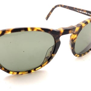 May include: Tortoiseshell-patterned sunglasses with green lenses. The frames have a classic cat-eye shape. The sunglasses are angled, showing the details of the frame and lenses.