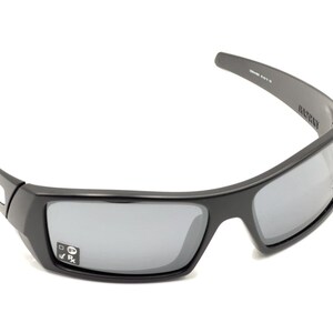 Puede incluir: Gafas de sol Oakley negras con lentes gris oscuro. Las gafas tienen un marco negro y un logotipo negro en el lateral.