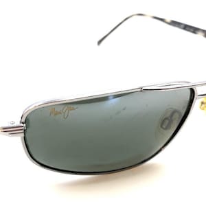 May include: Silver-framed sunglasses with green lenses. The sunglasses feature a double bridge and patterned temples. The brand name "Maui Jim" is visible on the lens. The design is a classic aviator style.