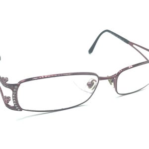 Pode incluir: Um par de óculos com armação de metal bordô. As armações têm formato retangular com lentes transparentes. As hastes são cinza escuro. O lado esquerdo da armação tem strass decorativos.