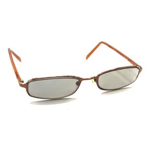 Könnte beinhalten: Rechteckige Sonnenbrille mit braunem Metallrahmen und getönten Gläsern. Die Bügel sind hellbraun. Die Brille ist vor einem weißen Hintergrund platziert.