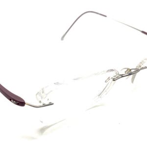 Pode incluir: Óculos sem aro com armação prateada e hastes bordô. As lentes são transparentes e os óculos têm um design minimalista. As hastes curvam-se suavemente.