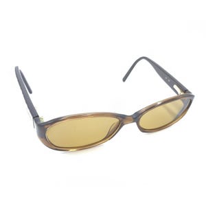 May include: Oval-shaped eyeglasses featuring a translucent brown frame and tinted amber lenses. The temples are dark brown with a metallic hinge detail connecting to the front frame. These glasses have a classic, rounded silhouette and curved earpieces.