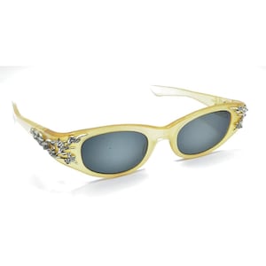 May include: Sunglasses with a translucent pale yellow frame and dark grey lenses. The outer corners of the frame feature silver-toned metal embellishments shaped like floral vines with small clear crystals embedded throughout the design.
