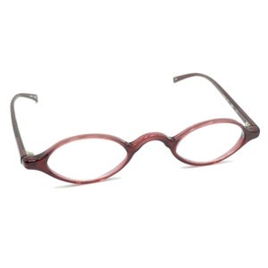 May include: A pair of eyeglasses with a translucent, deep reddish-purple frame. The lenses are oval-shaped, and the frame features a high bridge design with thin, matching temples that extend outward from the outer edges of the lens rims.