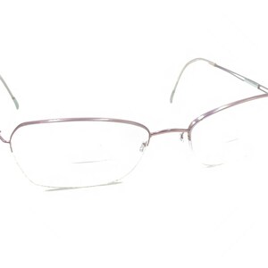 Puede incluir: Un par de gafas con una montura fina de metal morado claro. Las lentes transparentes tienen un diseño bifocal. Las varillas delgadas se extienden hasta los terminales. Las gafas están sobre un fondo blanco.