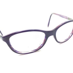 Puede incluir: Un par de gafas con montura morada oscura y lentes transparentes. La montura de ojo de gato tiene un sutil degradado rosa y morado en los bordes interiores. Las patillas son de un tono morado más claro.