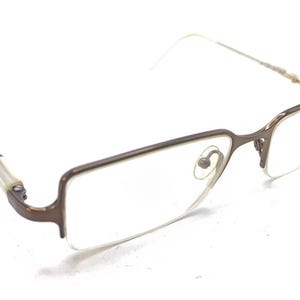 May include: Bronze-coloured rectangular-framed eyeglasses. The temples are a light colour, and the lenses are clear. The glasses have a modern design and are set against a white background.